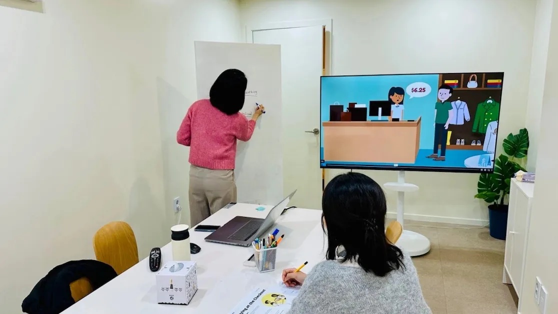 화이트보드에 쓰는 강사와 TV 화면의 영상 자료, 테이블에 앉아 수업을 듣는 수강생